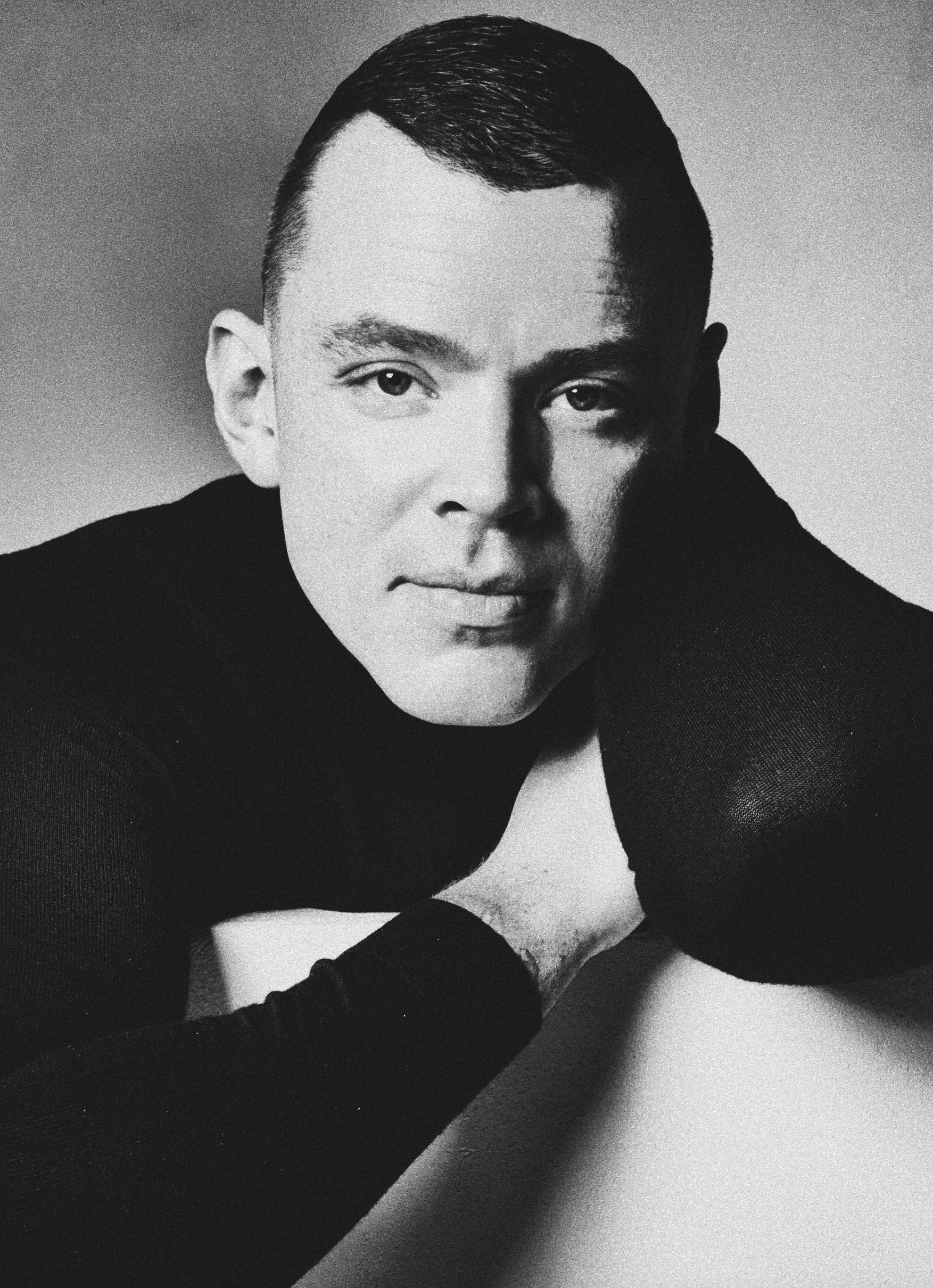 Black-and-white studio portrait of a man in a dark roll-neck, chin resting on hand