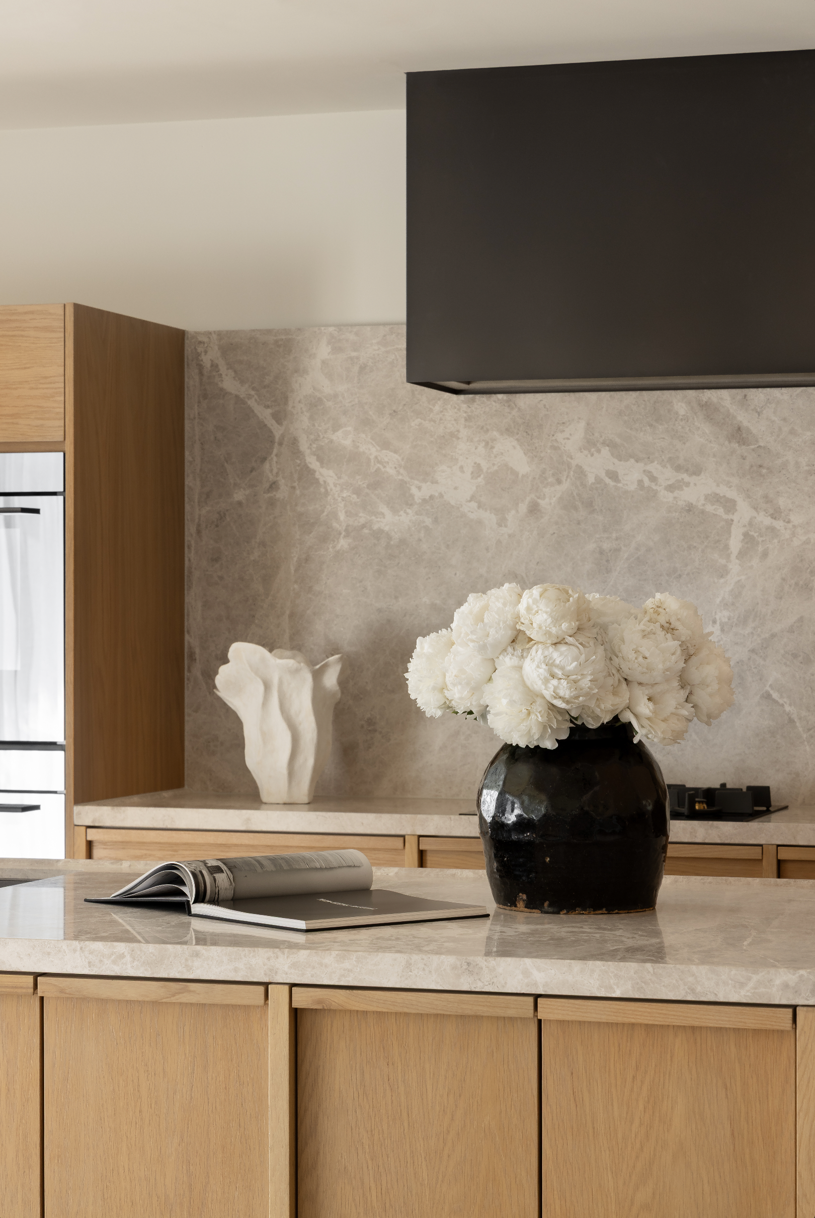 Stone benchtop and splashback with white peonies in a black vessel above timber cabinetry