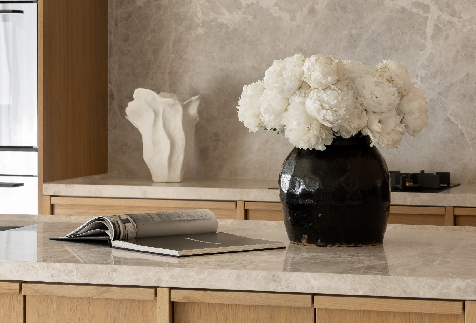 Stone benchtop and splashback with white peonies in a black vessel above timber cabinetry
