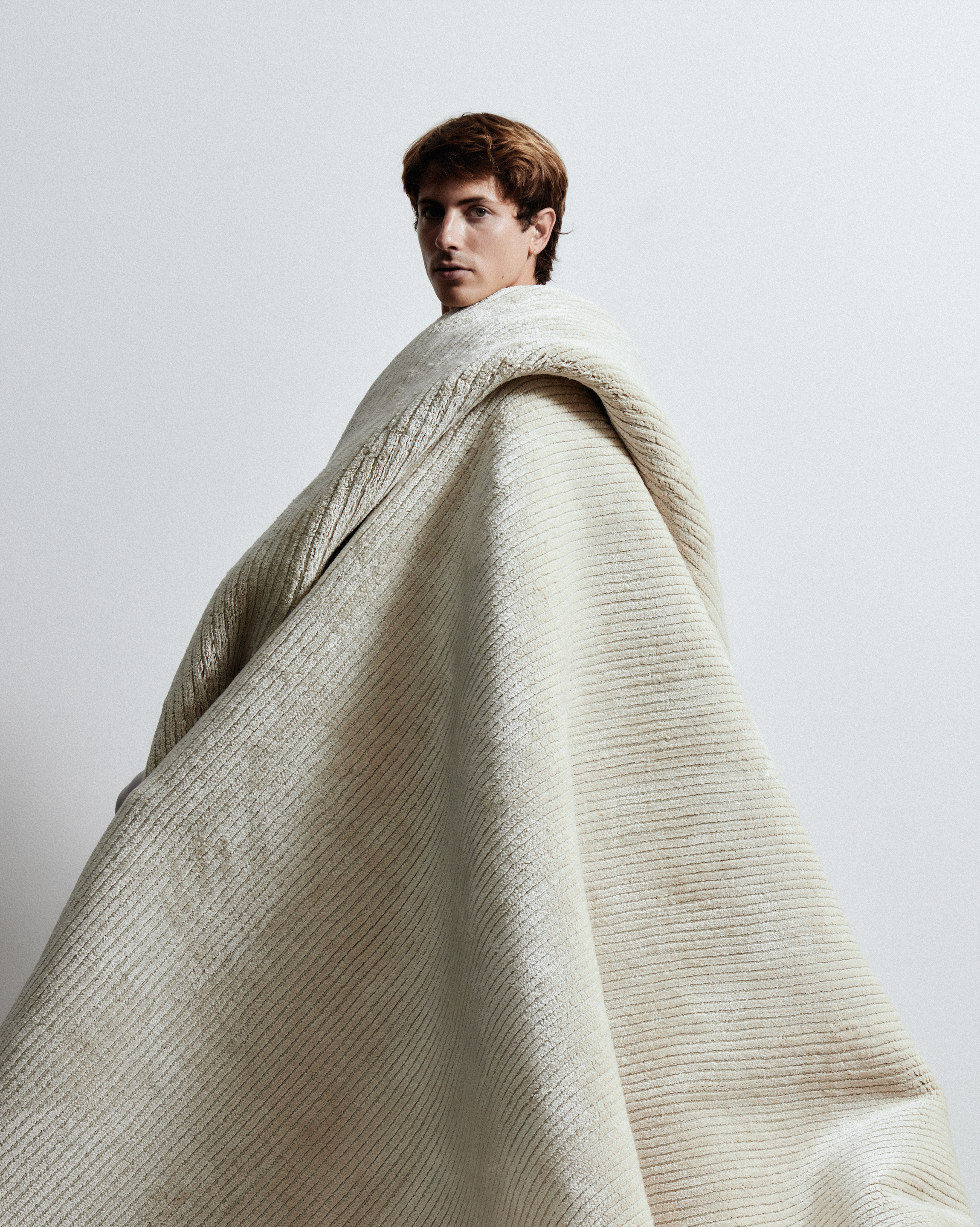 Model wrapped standing in a thick cream knit blanket against a bare, softly lit wall