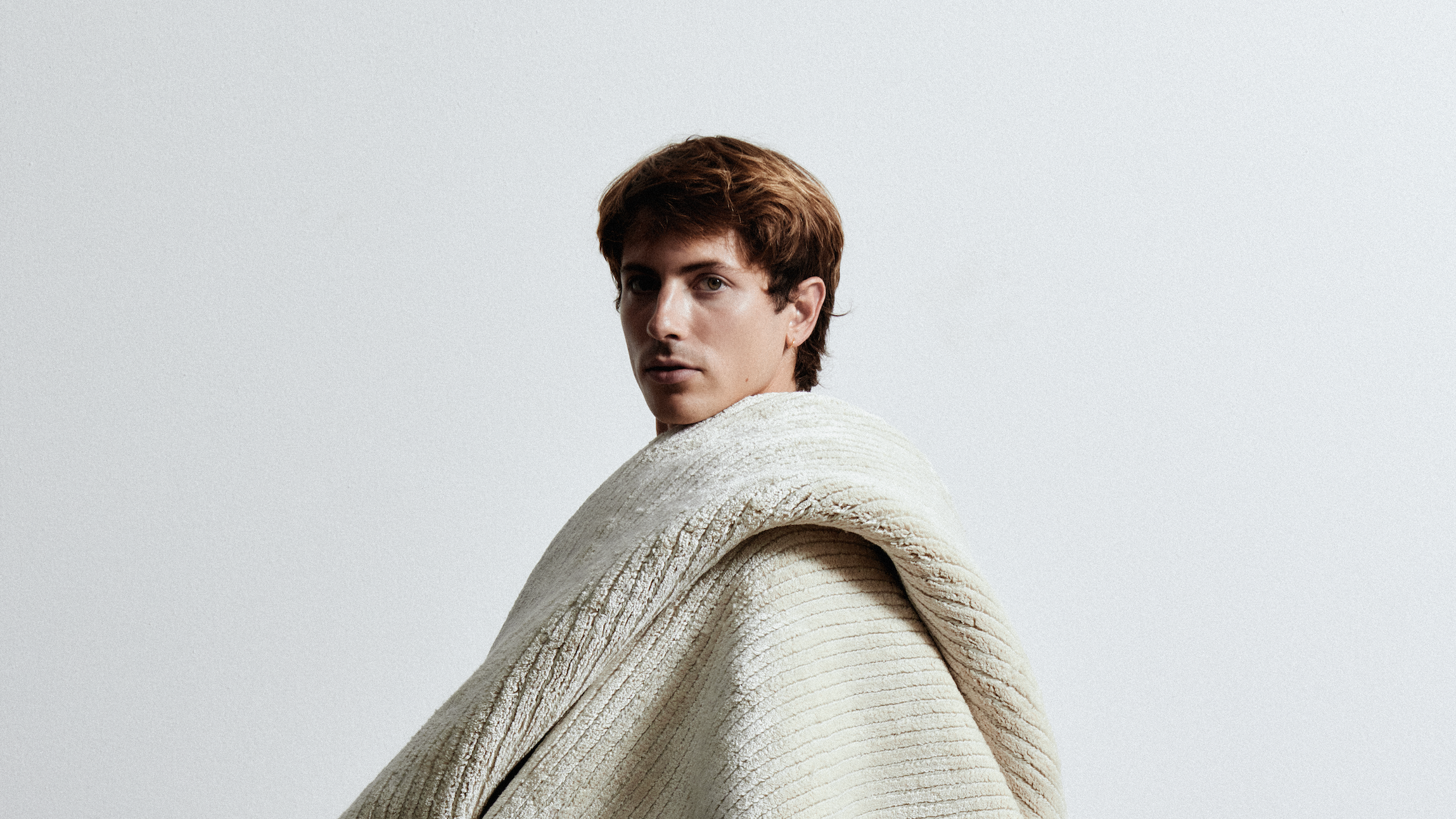 Model wrapped standing in a thick cream knit blanket against a bare, softly lit wall