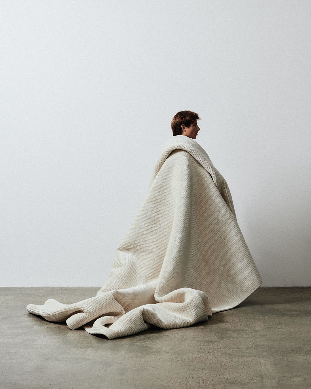 Seated figure cocooned on the floor in a cream knit blanket against a bare wall