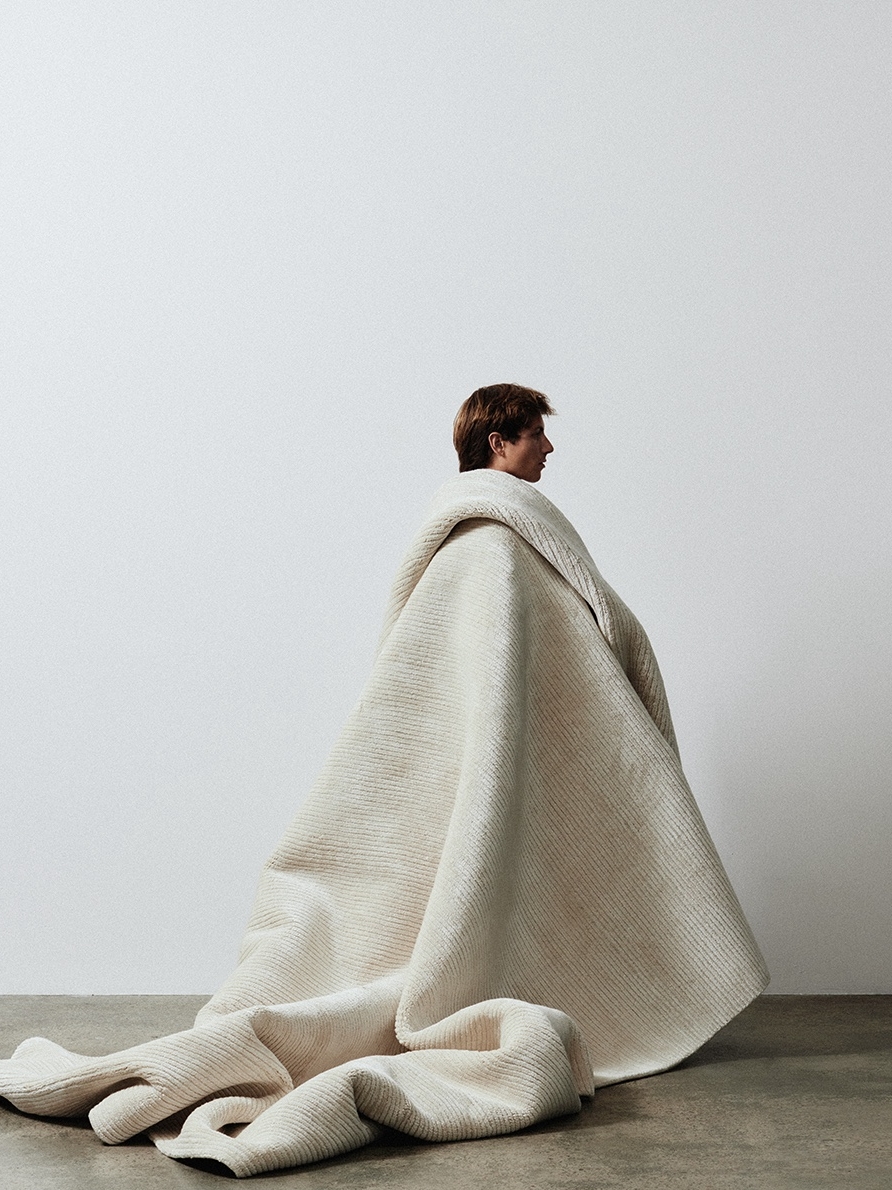 Seated figure cocooned on the floor in a cream knit blanket against a bare wall
