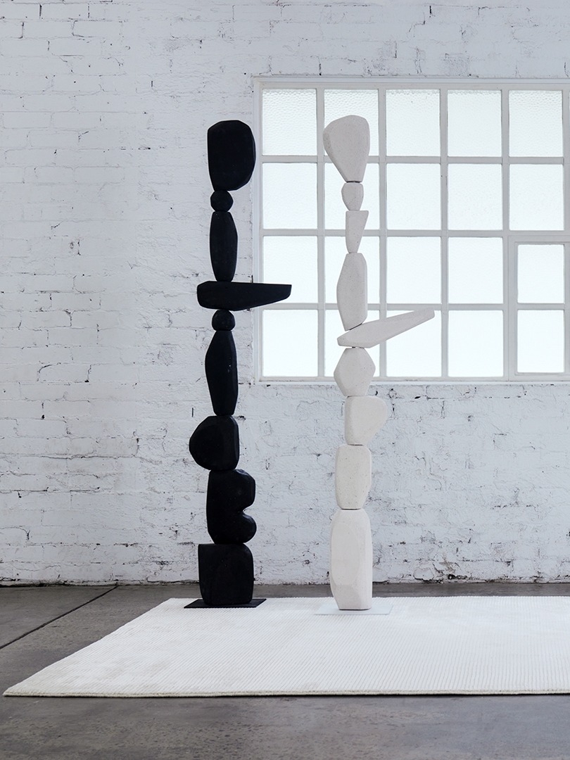 Two stacked totem sculptures, one black and one pale, before a gridded studio window
