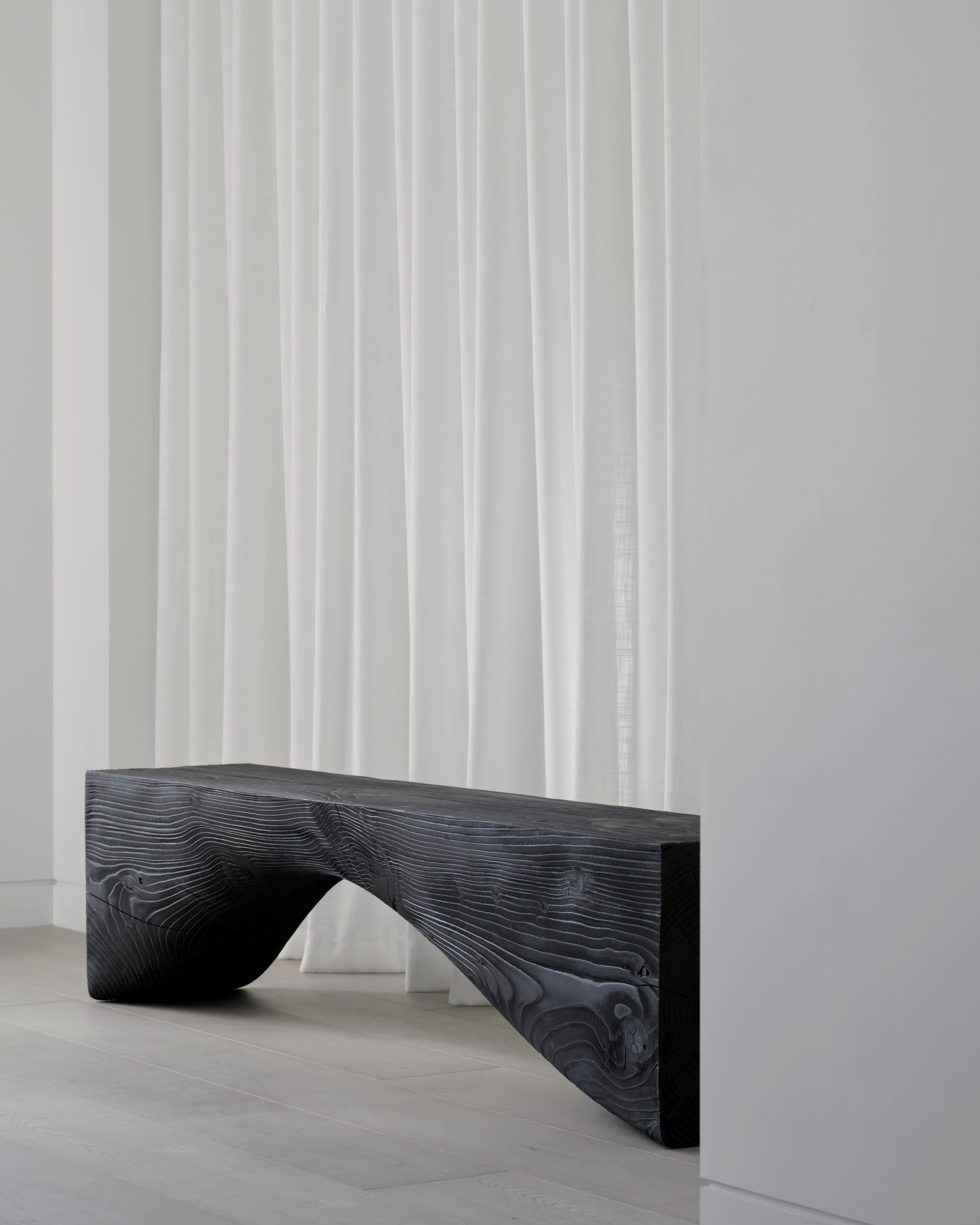 Charred timber bench curving like a wave against sheer floor-length curtains