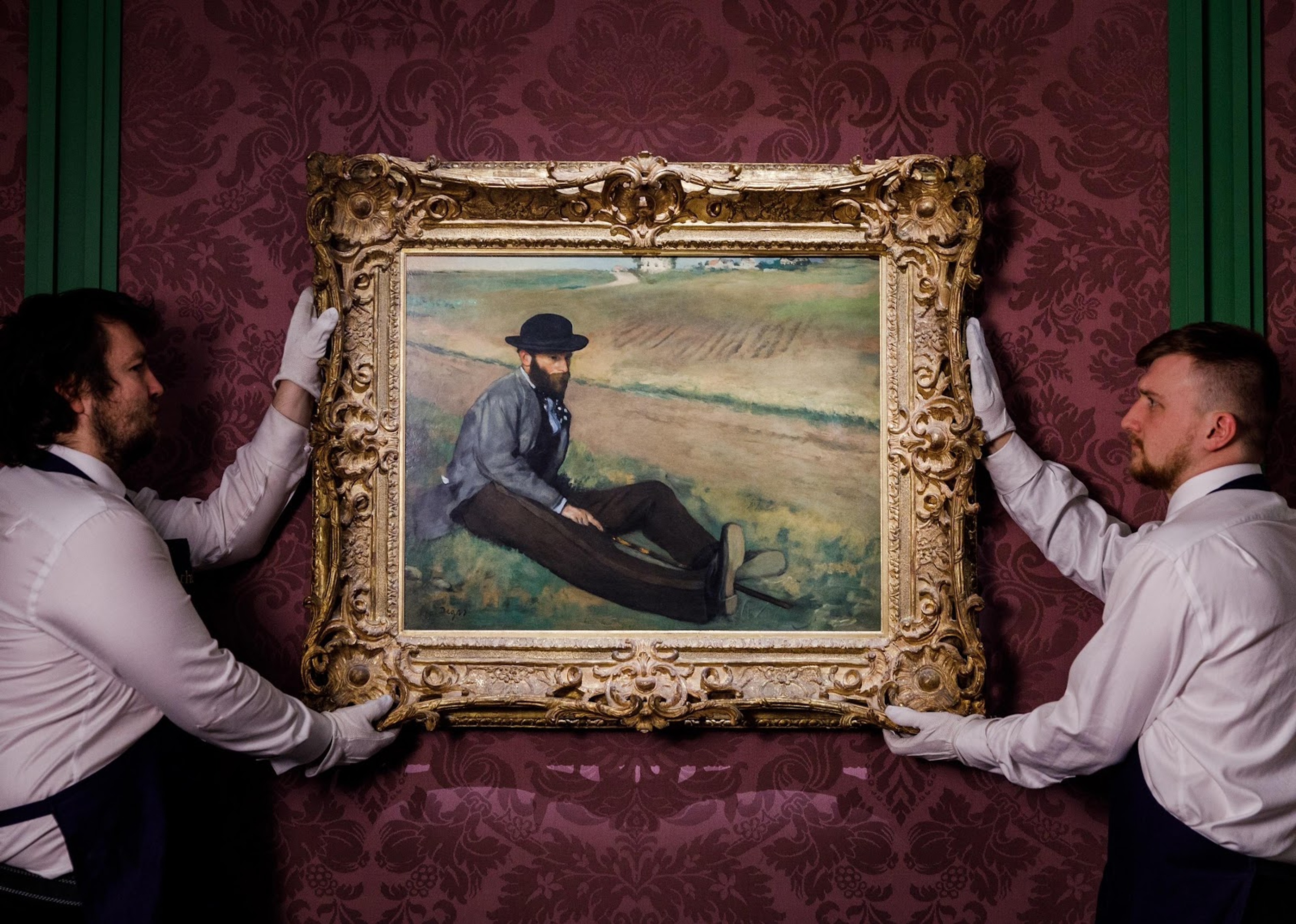 White-gloved handlers hanging a gilt-framed Impressionist painting on a burgundy damask wall