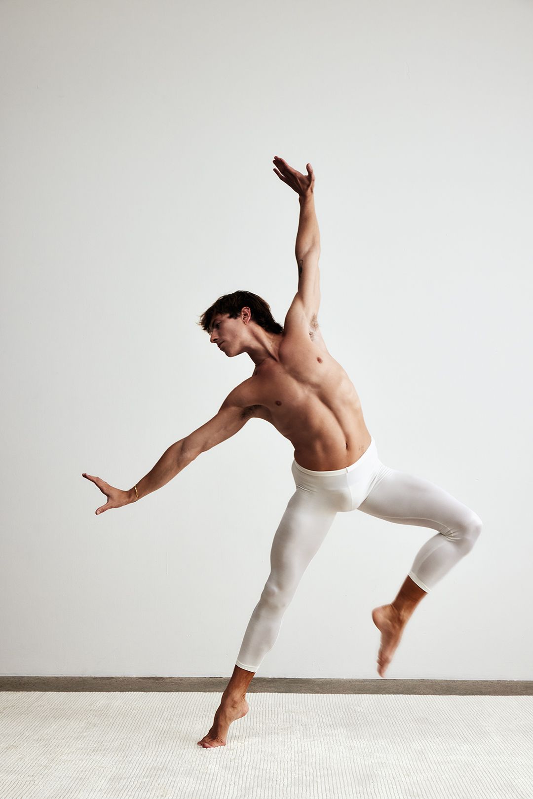Male dancer captured mid-arabesque in white tights against a pale studio backdrop
