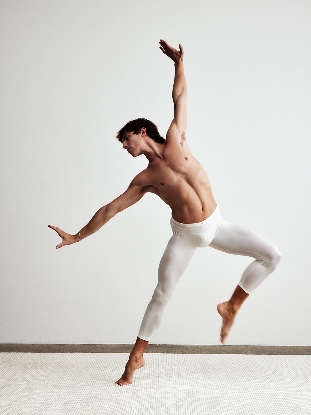 Male dancer captured mid-arabesque in white tights against a pale studio backdrop