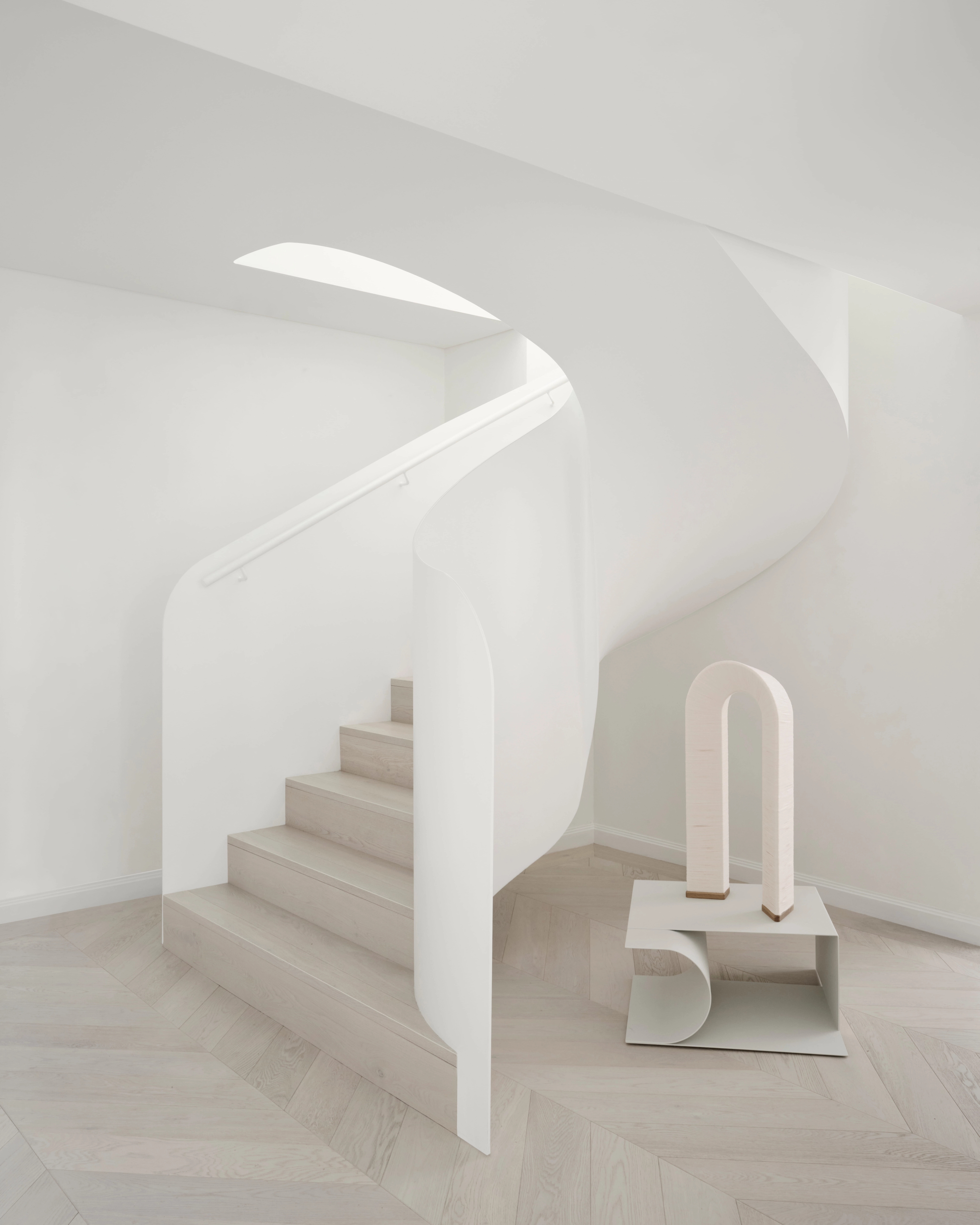 Sculpted white staircase curving above herringbone timber beside a minimalist arch object