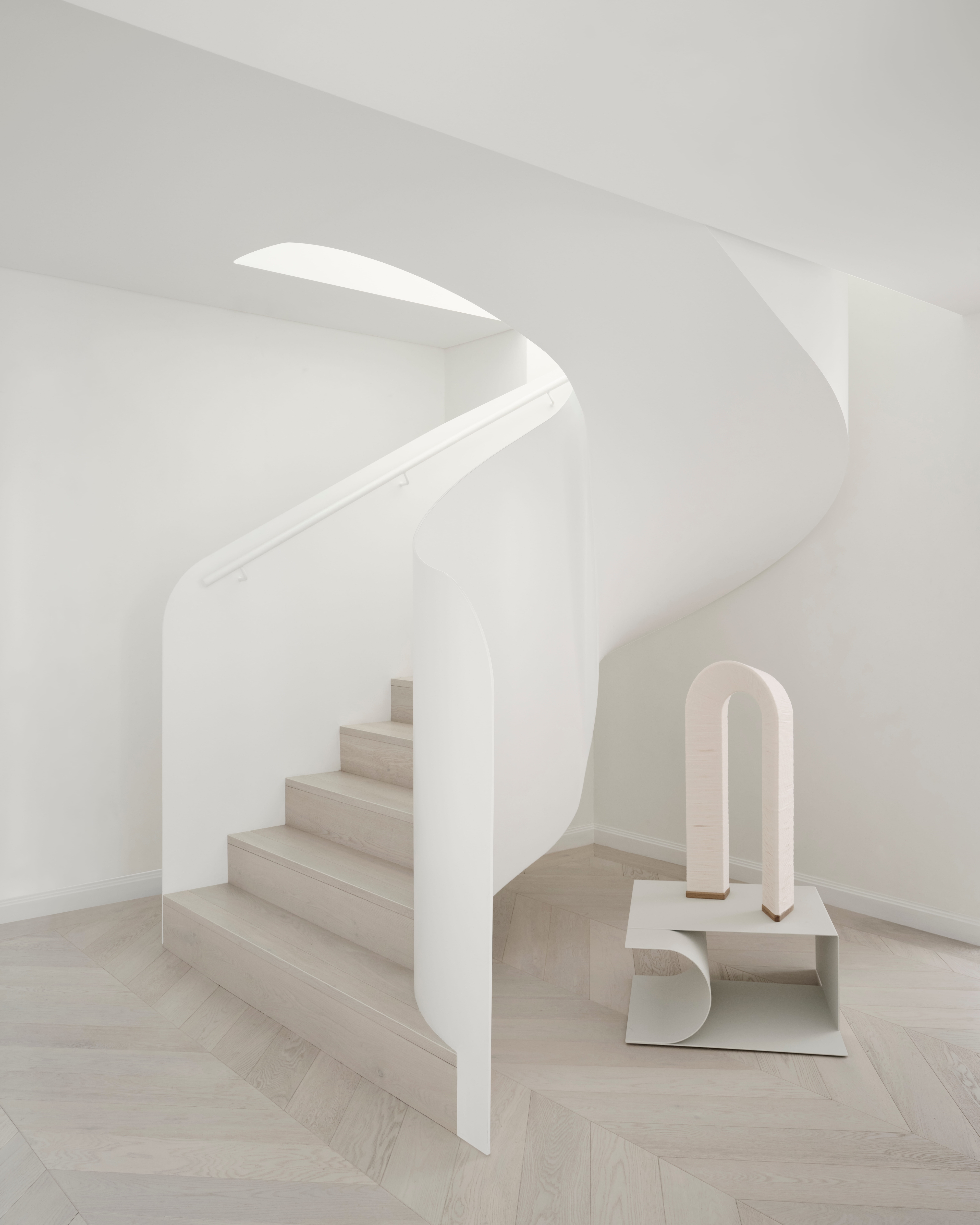 Sculpted white staircase curving above herringbone timber beside a minimalist arch object