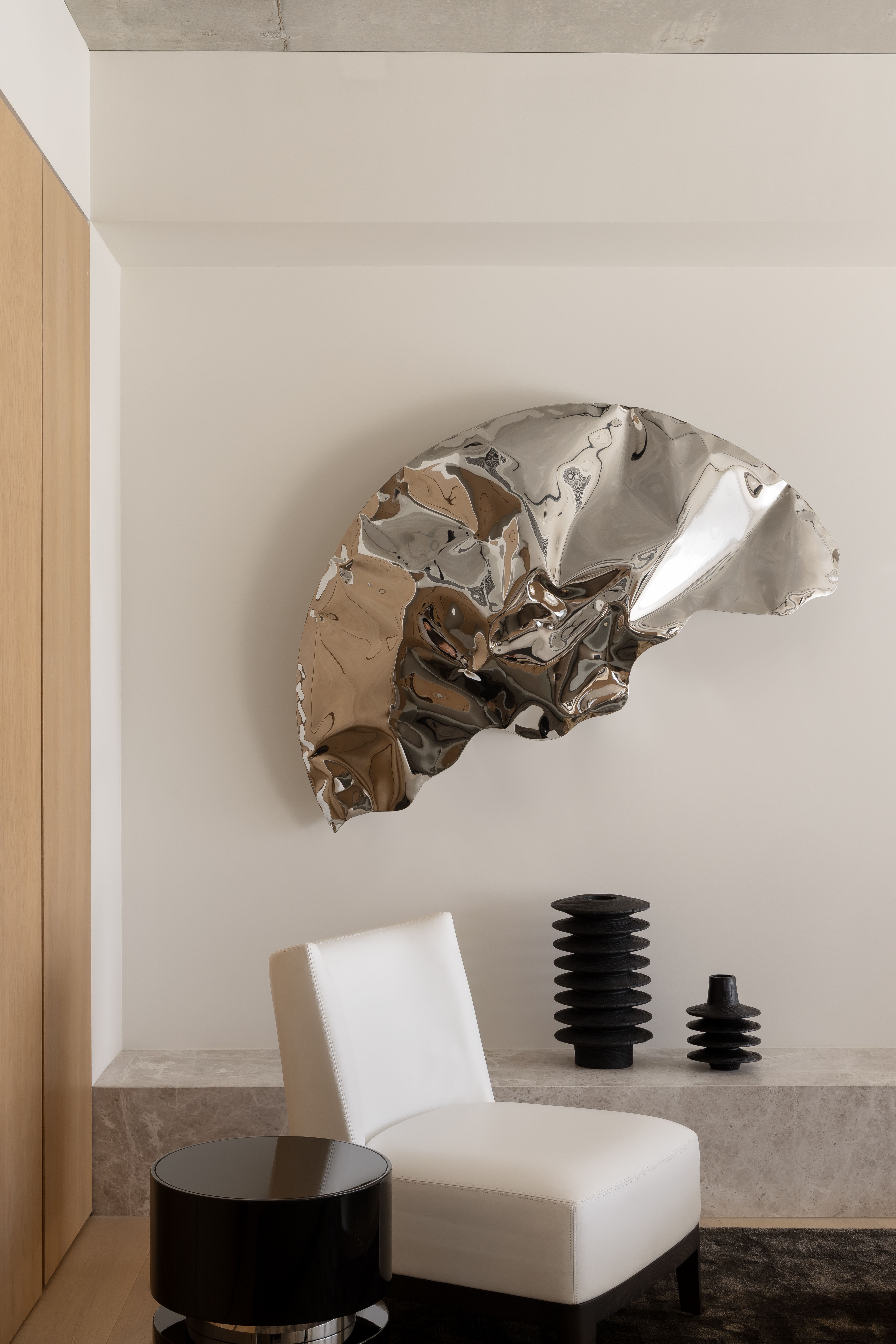 Crumpled chrome wall sculpture above a white chair and black totem objects
