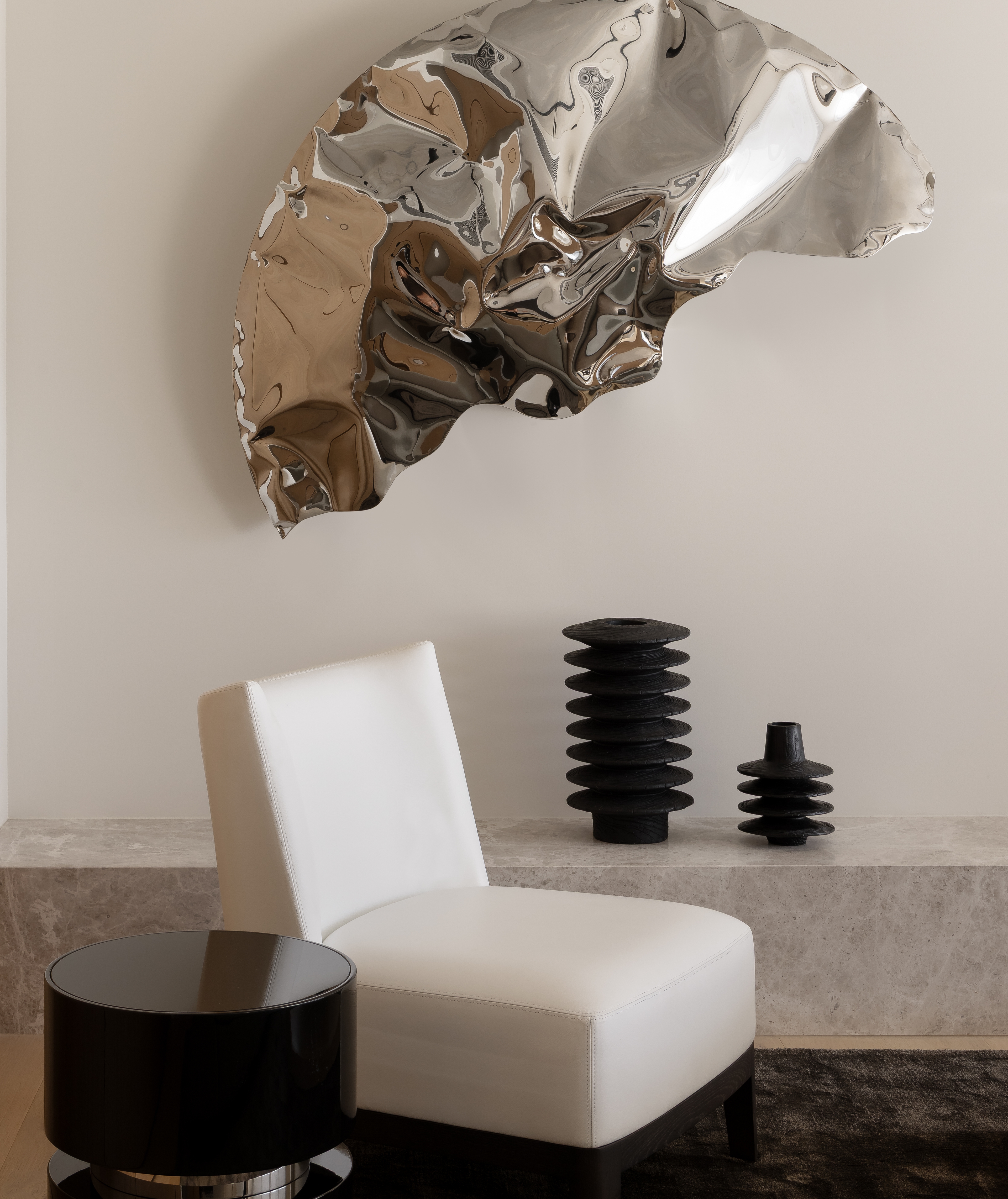 Crumpled chrome wall sculpture above a white chair and black totem objects