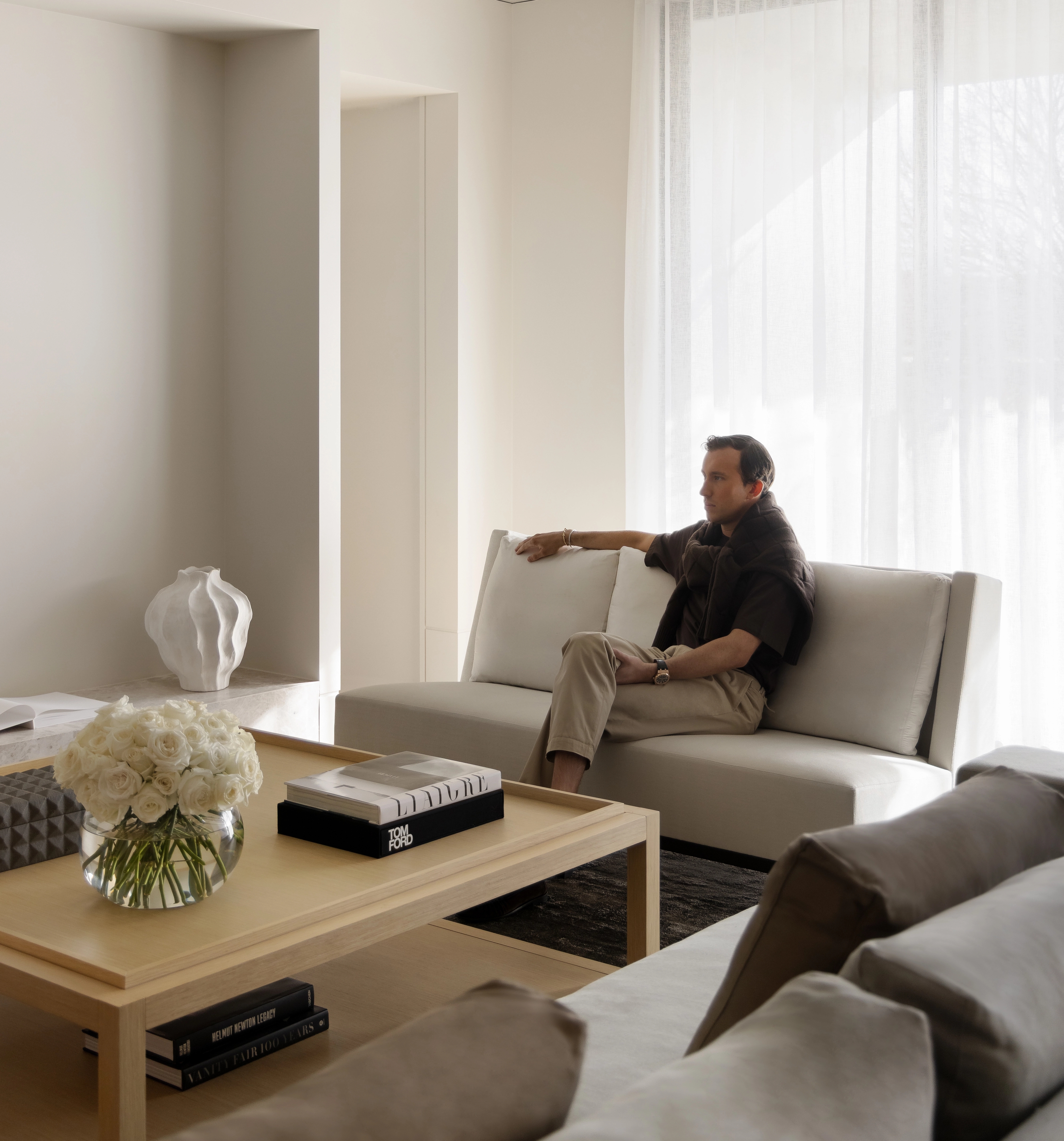 Relaxed figure on a taupe sofa beside a low table stacked with books and white blooms