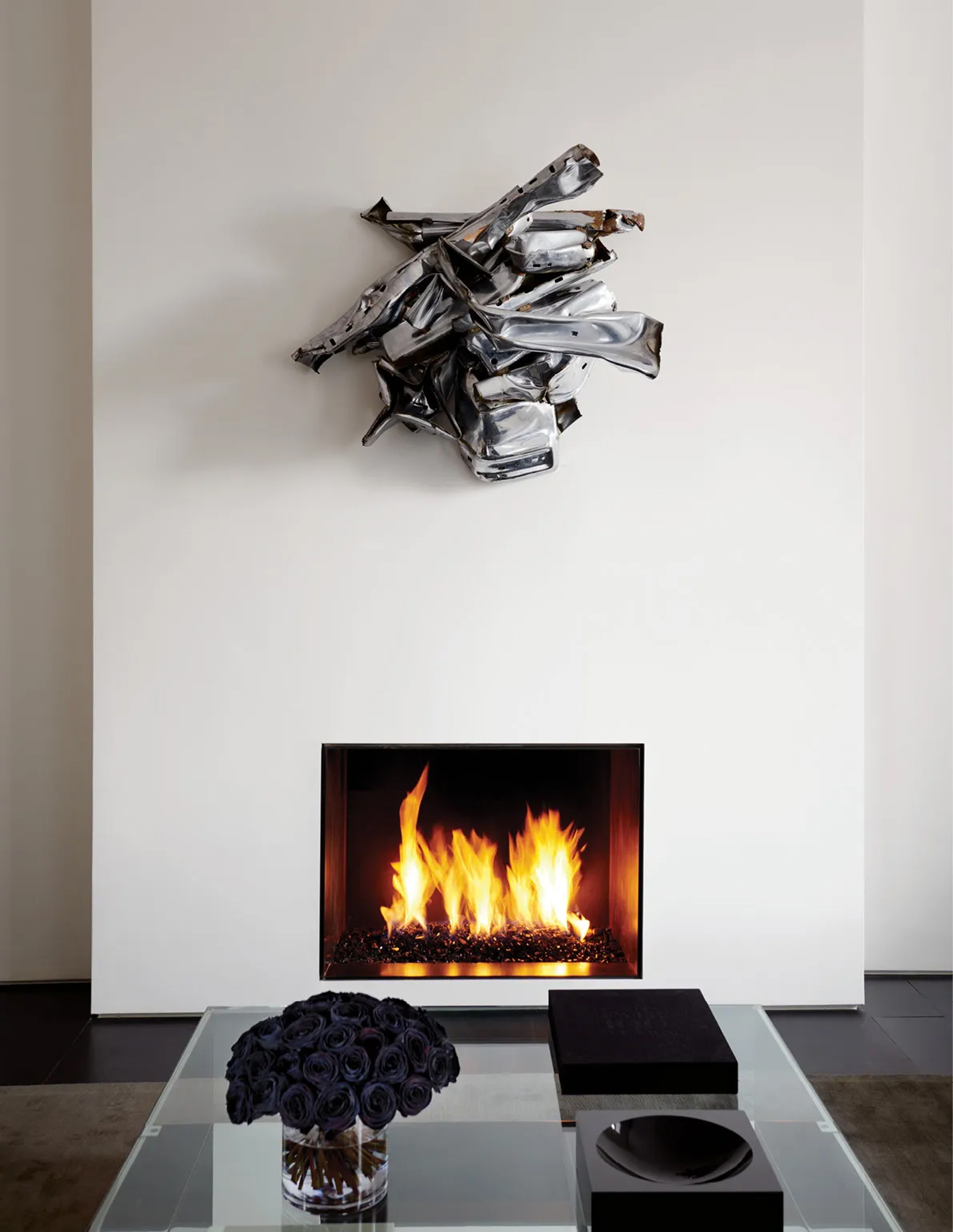 Silver crumpled sculpture above a lit fireplace with dark roses on glass