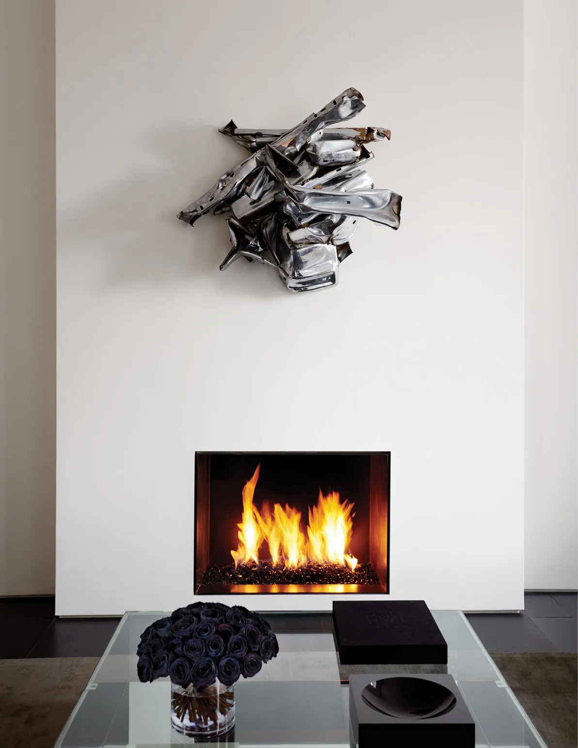 Silver crumpled sculpture above a lit fireplace with dark roses on glass